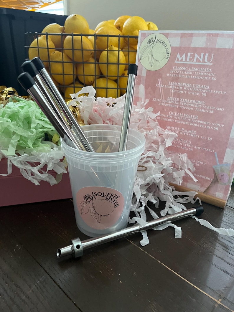 K&ouml;nnte beinhalten: Nahaufnahme eines durchsichtigen Plastikbechers mit einem rosa-wei&szlig;en Logo, auf dem "Squeeze Sister Lemonade & Tea" steht. Der Becher ist mit bunten Papierschnitzeln und vier wiederverwendbaren Edelstahlstrohhalmen gef&uuml;llt. Ein Edelstahlstrohhalm mit schwarzer Spitze liegt auf einem Holztisch.