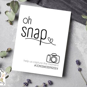 May include: White card with black text that says "Oh snap" with a heart and a camera icon. The text below the image says "Help us capture the moment #JONESWEDDING2024".