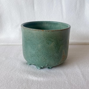 May include: A green ceramic planter with a textured finish and small feet. The planter has drainage holes in the bottom.