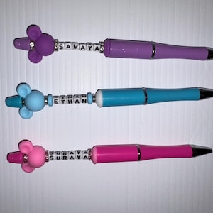 May include: Three personalized pens with colorful animal toppers and beads spelling out the names "Samaya", "Ethan", and "Suraya".