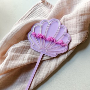 May include: A lavender seashell-shaped cake topper with the name "emma" in pink script. The seashell has a glittery silver outline and a matching lavender stem. The topper is designed for cakes and other desserts.