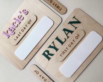 May include: Two wooden signs with blank spaces for writing in. One sign says "Rylan" in green letters and "First Day Of" in brown letters. The other sign says "Kacie's" in pink letters and "Last Day Of" in brown letters.