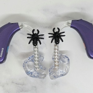 May include: A pair of purple Phonak hearing aids with clear ear moulds containing blue glitter. Each device features a black spider charm attached to a silver coiled tube that connects the hearing aid body to the ear mould piece.