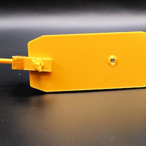 Op de afbeelding: Een geel 3D-geprint object van plastic met een kleine schroef en een hefboomarm. Het object is ontworpen om te dienen als standaard voor een klein object.