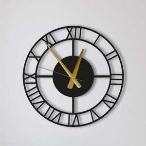May include: A large, round black metal wall clock with gold-colored hands and Roman numerals. The clock is mounted on a white wall above a beige sofa. The clock's face has a black center.