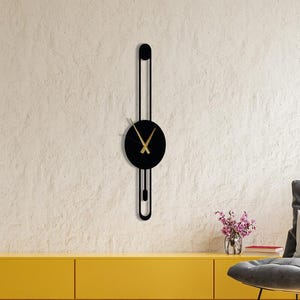 May include: A modern, black wall clock with gold-colored hands and a circular face. The clock has a unique, elongated design with a vertical bar and a curved bottom. The clock is mounted on a textured, off-white wall.
