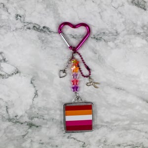 May include: A pink heart-shaped carabiner clip with a keychain featuring a square pendant with the lesbian pride flag colours. The keychain has star-shaped beads in rainbow colours, a heart charm, and small scissors.