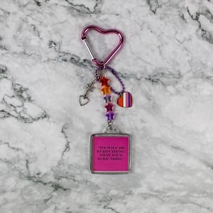 May include: A pink heart-shaped carabiner keychain with a square charm. The charm has a pink background with the text "WE WILL BE EVERYTHING THAT WE'D EVER NEED." The keychain has star and heart charms.