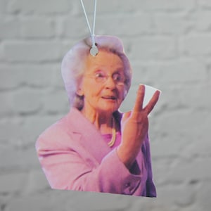 May include: A pink car air freshener featuring a woman with white hair and a pink jacket. She is smiling and holding up two fingers.
