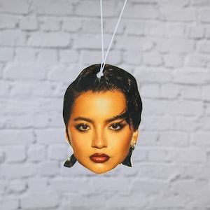 May include: A car air freshener in the shape of a woman's face, featuring dark hair and red lipstick. It hangs from a white string, set against a white brick wall.
