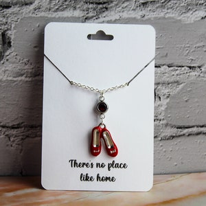 May include: A silver necklace with a red ruby charm and a pair of red shoes. The charm is attached to the necklace with a silver chain. The necklace is on a white card with the text "There's no place like home".