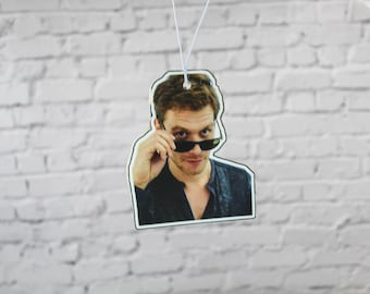 May include: Air freshener featuring a man with brown hair and sunglasses, looking over the top of the glasses. The air freshener is hanging from a white string against a white brick wall background.