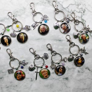 May include: Several silver keyrings, each with a round pendant displaying a portrait. The keyrings have a clasp, a ring, and decorative charms such as crowns, swords, and beads. The pendants have a glossy finish.