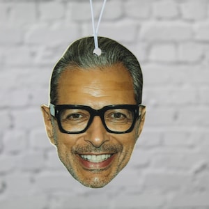 May include: A novelty air freshener with a smiling face wearing black-framed glasses. The image is a close-up of a person's face, with a white string for hanging. The background is a white brick wall.