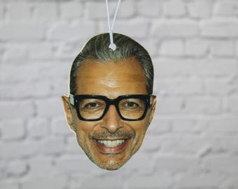 May include: A decorative air freshener featuring a smiling face with black-framed glasses. The image is a close-up of a person's face, with a white string for hanging. The background is a white brick wall.