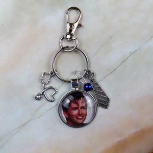 May include: A silver-toned metal keyring featuring a circular pendant with a photographic portrait of a man, a small silver stethoscope charm, a silver tie charm with diagonal stripes, and a single small round dark blue bead on a jump ring.