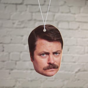 May include: A car air freshener featuring a printed image of a man with a moustache. The man is looking directly at the viewer.