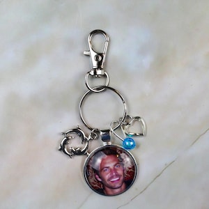 May include: A silver-toned metal keychain featuring a circular pendant with a photographic portrait of a man. The keychain includes a lobster clasp, a split ring, a charm of two jumping dolphins, a small blue bead, and a silver-toned heart charm.