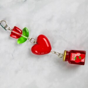 May include: A charm featuring a red rose, green leaves, a red heart, and a miniature jar of strawberry jam. The charm is connected by silver-coloured chains and a black cord.
