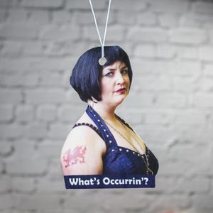 Peut inclure: Une femme aux cheveux noirs courts porte un haut bleu fonc&eacute; sans manches avec un collier argent&eacute;. Elle a un tatouage de dragon rouge sur l'&eacute;paule gauche. Le texte "What's Occurrin'?" est imprim&eacute; sur une banni&egrave;re blanche en dessous d'elle.