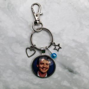 Könnte beinhalten: Silberfarbener Schlüsselanhänger mit einem runden Anhänger, der eine lächelnde Person zeigt. Der Schlüsselanhänger enthält einen Herzanhänger, einen Sternanhänger und eine blaue Perle. Der Hintergrund ist ein hellgrauer Marmor.