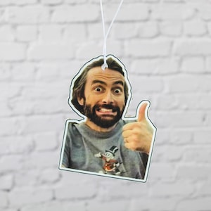 May include: Rectangular air freshener featuring a man with a beard giving a thumbs up. The air freshener has a white string for hanging. The man is wearing a grey t-shirt with a cartoon graphic. The background is a white brick wall.