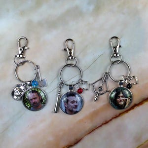 May include: Three silver-toned metal keychains featuring circular glass cabochons with portraits of characters. Each keychain includes a lobster clasp, a split ring, and small charms: a hat and axe, a baseball bat, and a crossbow with a heart.
