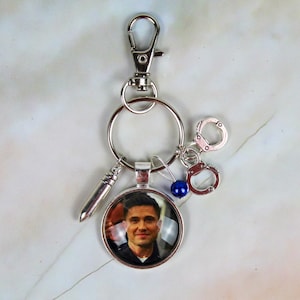 Peut inclure: Porte-clés argenté avec un pendentif rond représentant le portrait d'un homme. Le porte-clés comprend un charme de balle, de menottes miniatures et une perle bleue. Un mousqueton et un porte-clés sont également présents.