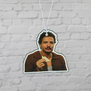 May include: An air freshener featuring a color image of a man with dark hair and a mustache, wearing a brown sweater and holding a piece of food. The air freshener is hanging from a white string against a white brick wall.