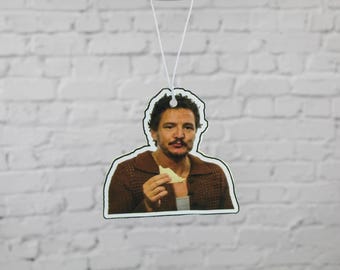 Puede incluir: Un ambientador con una imagen en color de un hombre con cabello oscuro y bigote, que lleva un suéter marrón y sostiene un trozo de comida. El ambientador cuelga de un hilo blanco contra una pared de ladrillos blancos.