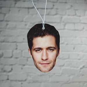 May include: A car air freshener featuring a close-up image of a man's face. The man has short brown hair and is looking directly at the camera.