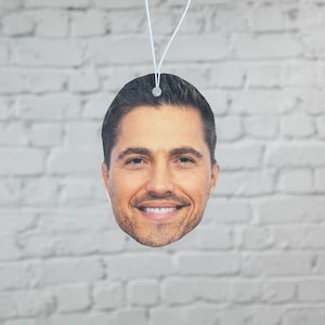 May include: A car air freshener shaped like a smiling man's face. The air freshener is a light tan color with a white string for hanging. The background is a white brick wall.