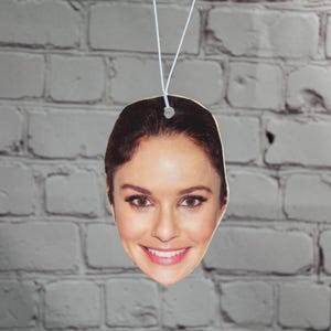 Puede incluir: Un ambientador de coche con la cara de una mujer con cabello casta&ntilde;o. El ambientador es blanco y tiene una cuerda para colgar.