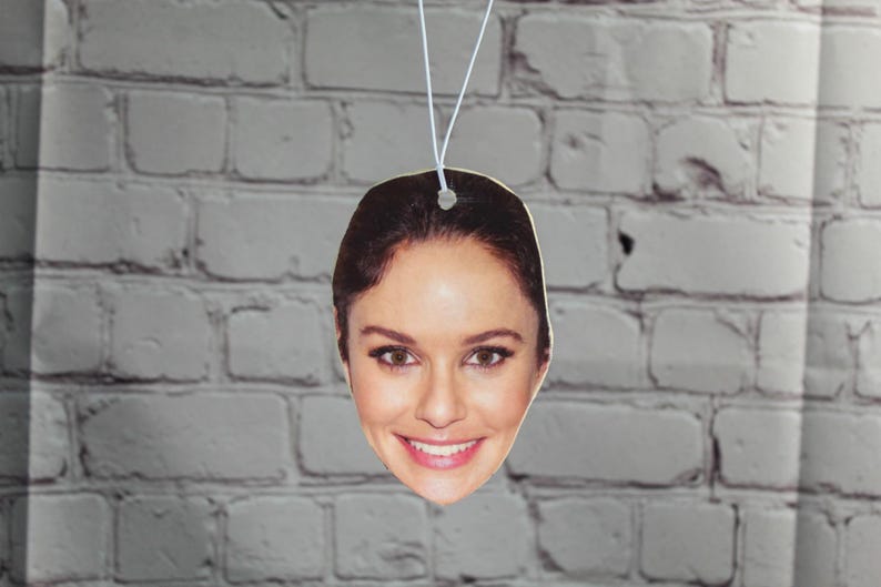 Puede incluir: Un ambientador de coche con la cara de una mujer con cabello casta&ntilde;o. El ambientador es blanco y tiene una cuerda para colgar.