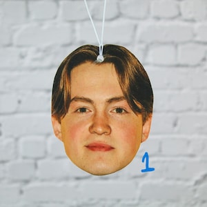 May include: Oval air freshener with a young person's face, light brown hair and fair skin. A white string is attached for hanging. The background is a white brick wall. A blue number 1 is in the lower right corner.