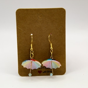 Puede incluir: Un par de pendientes de oro con un dije de paraguas colorido y una cuenta azul claro colgando de cada uno. Los pendientes se muestran en una tarjeta marrón con el texto "Handmade with Love".