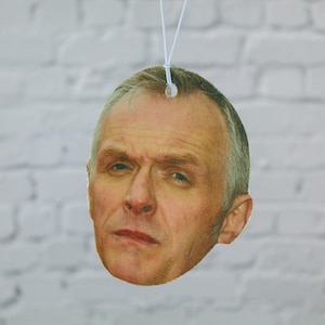 May include: Oval-shaped air freshener featuring a man's face, suspended by a white string. The man has short gray hair and a serious expression. The background is a white brick wall.