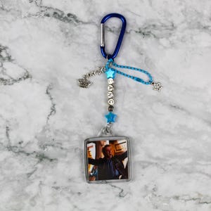 Può includere: Un portachiavi con moschettone blu con un ciondolo quadrato trasparente con la foto di un uomo. Il portachiavi ha stelle blu e perline, e la parola "DANCE" scritta con perline.