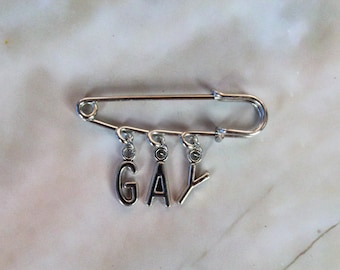Pode incluir: Um alfinete de segurança prateado com quatro pingentes que soletram a palavra "GAY".