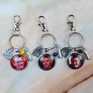 May include: Three silver-toned keychains, each featuring a circular pendant with a portrait of a man, a silver flame charm, a small silver fire extinguisher charm, and a single coloured bead in yellow, blue, or black attached to a lobster clasp.