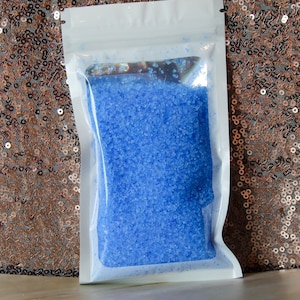 Puede incluir: Una bolsa de plástico transparente llena de sales de baño de color azul brillante. La bolsa está sellada y lista para usar.
