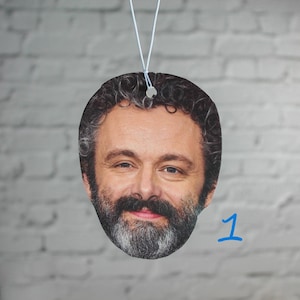 May include: A round car air freshener featuring a photo of a man with a beard and curly hair. The air freshener is hanging from a string.