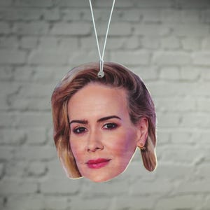 Puede incluir: Un ambientador de coche con la cara de una mujer con el pelo rubio. El ambientador es blanco y tiene una cuerda para colgar.