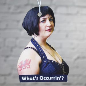 Peut inclure: Une femme aux cheveux noirs courts portant un haut bleu fonc&eacute; sans manches avec un collier en cha&icirc;ne argent&eacute;e et un tatouage de dragon rouge sur l'&eacute;paule gauche. Le texte "What's Occurrin'" est imprim&eacute; sur une banni&egrave;re bleue en dessous d'elle.