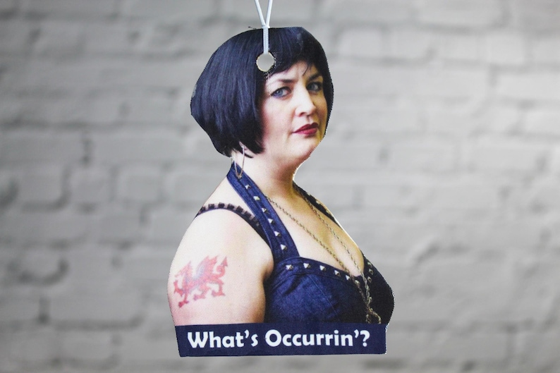 Peut inclure: Une femme aux cheveux noirs courts portant un haut bleu fonc&eacute; sans manches avec un collier en cha&icirc;ne argent&eacute;e et un tatouage de dragon rouge sur l'&eacute;paule gauche. Le texte "What's Occurrin'" est imprim&eacute; sur une banni&egrave;re bleue en dessous d'elle.