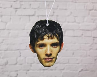 May include: A printed air freshener featuring a man's face with dark hair and light skin. The air freshener is hanging by a white string against a white brick wall background.