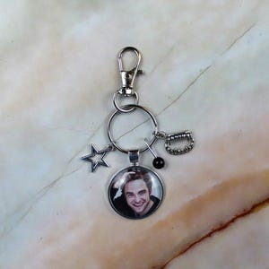 Op de afbeelding: Een zilverkleurige metalen sleutelhanger met een ronde hanger met een fotografisch portret van een man, een stervormige bedel, een kleine zwarte kraal aan een speld en een bedel in de vorm van vampiertanden aan een ring.