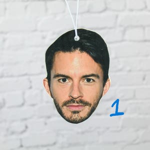 May include: A face-shaped air freshener featuring a man's portrait. The image is printed on a light-colored, oval-shaped piece of material, with a white string for hanging. The background is a white brick wall, and the number "1" is written in blue.