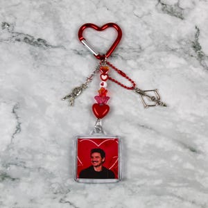May include: A red heart-shaped carabiner clip with a keychain featuring a photo of a person. The keychain has red and pink beads, a heart charm, and silver-colored charms. The background is a marble surface.