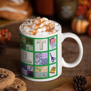 May include: White ceramic mug with a green and white patchwork design featuring the letters "LT" and sewing-related imagery. The mug is filled with a creamy beverage topped with whipped cream and cinnamon.